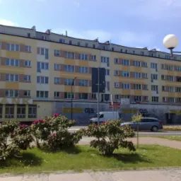 Wielopiętrowy budynek mieszkalny w trakcie remontu elewacji, widoczne rusztowania i siatki ochronne, zieleń miejska w pierwszym planie.